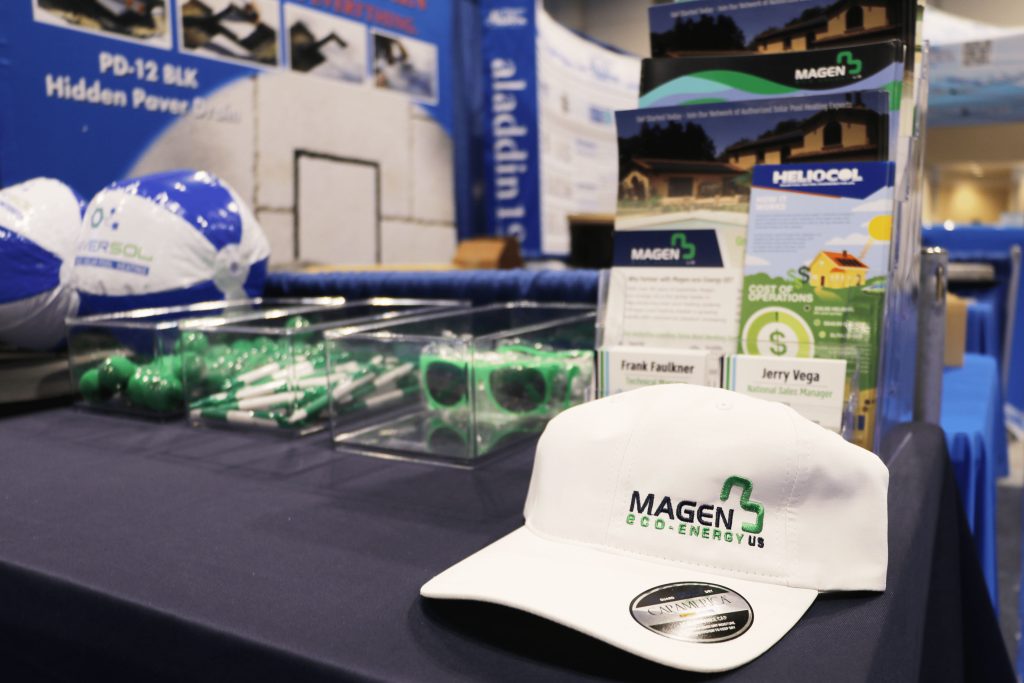 Magen eco-Energy US trade show booth display featuring branded marketing materials, white promotional hat, green sunglasses, pens, stress balls, and Heliocol product brochures, with signage for National Sales Manager Jerry Vega.